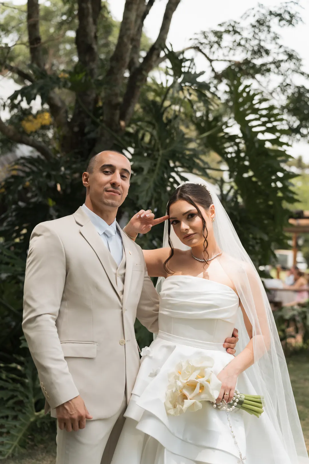 Noivos de bege posando no jardim, ela com copos-de-leite