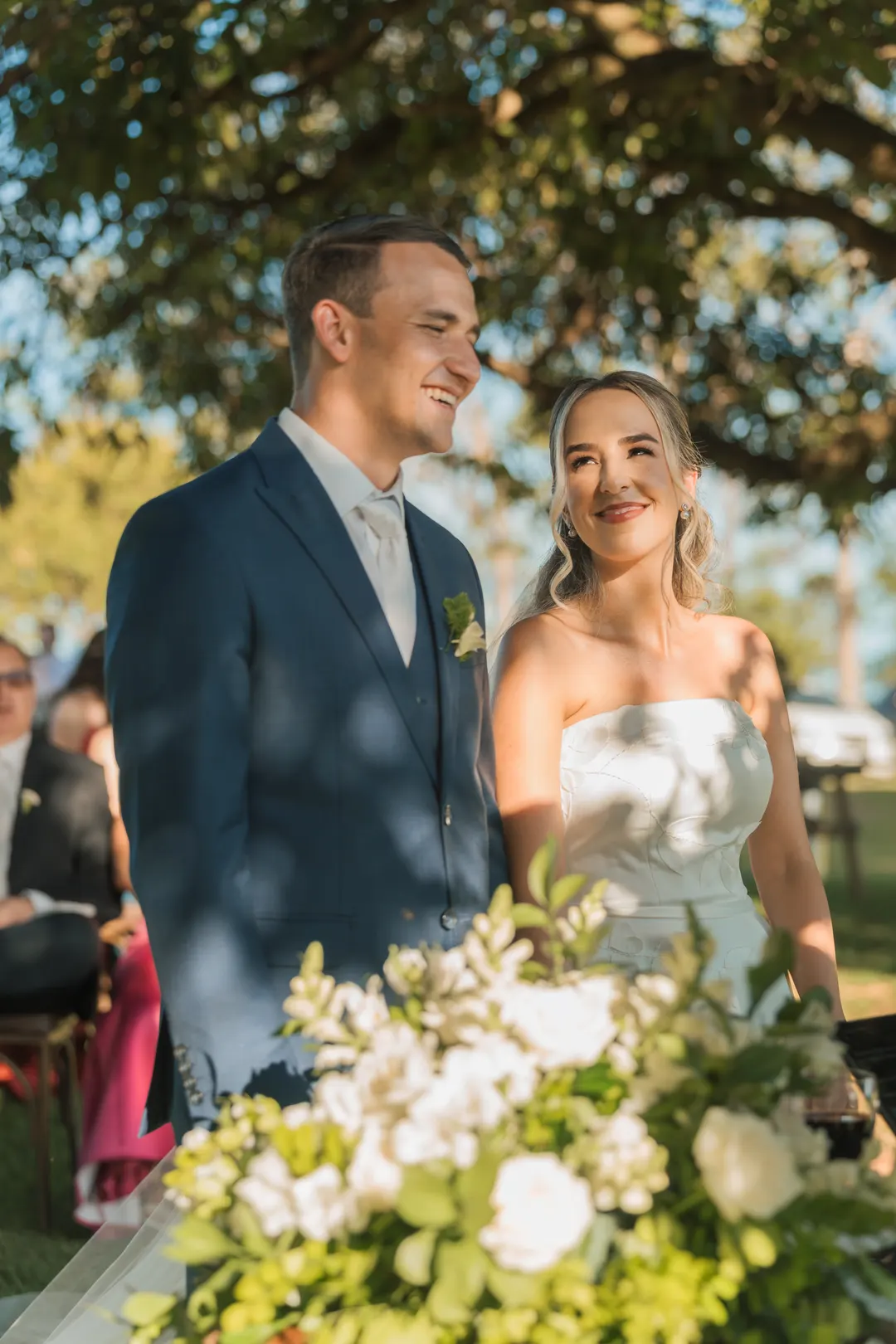 Noivos sorrindo no altar ao ar livre, com flores brancas à frente