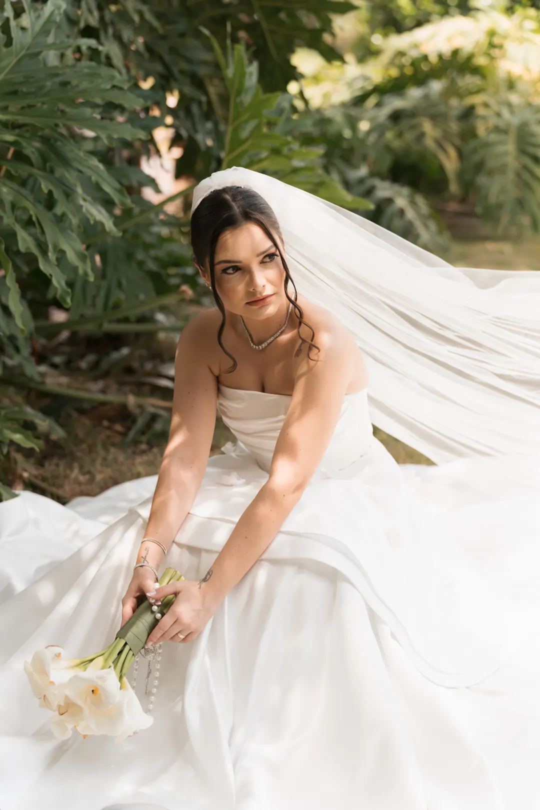 Noiva sentada no jardim com o véu ao vento, segurando o buquê