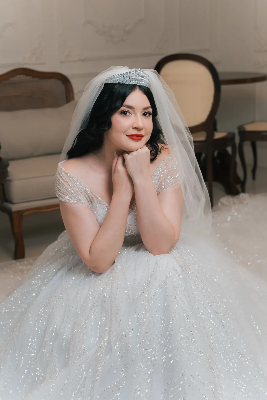 Noiva com tiara e vestido de brilho, mãos no rosto, sorrindo