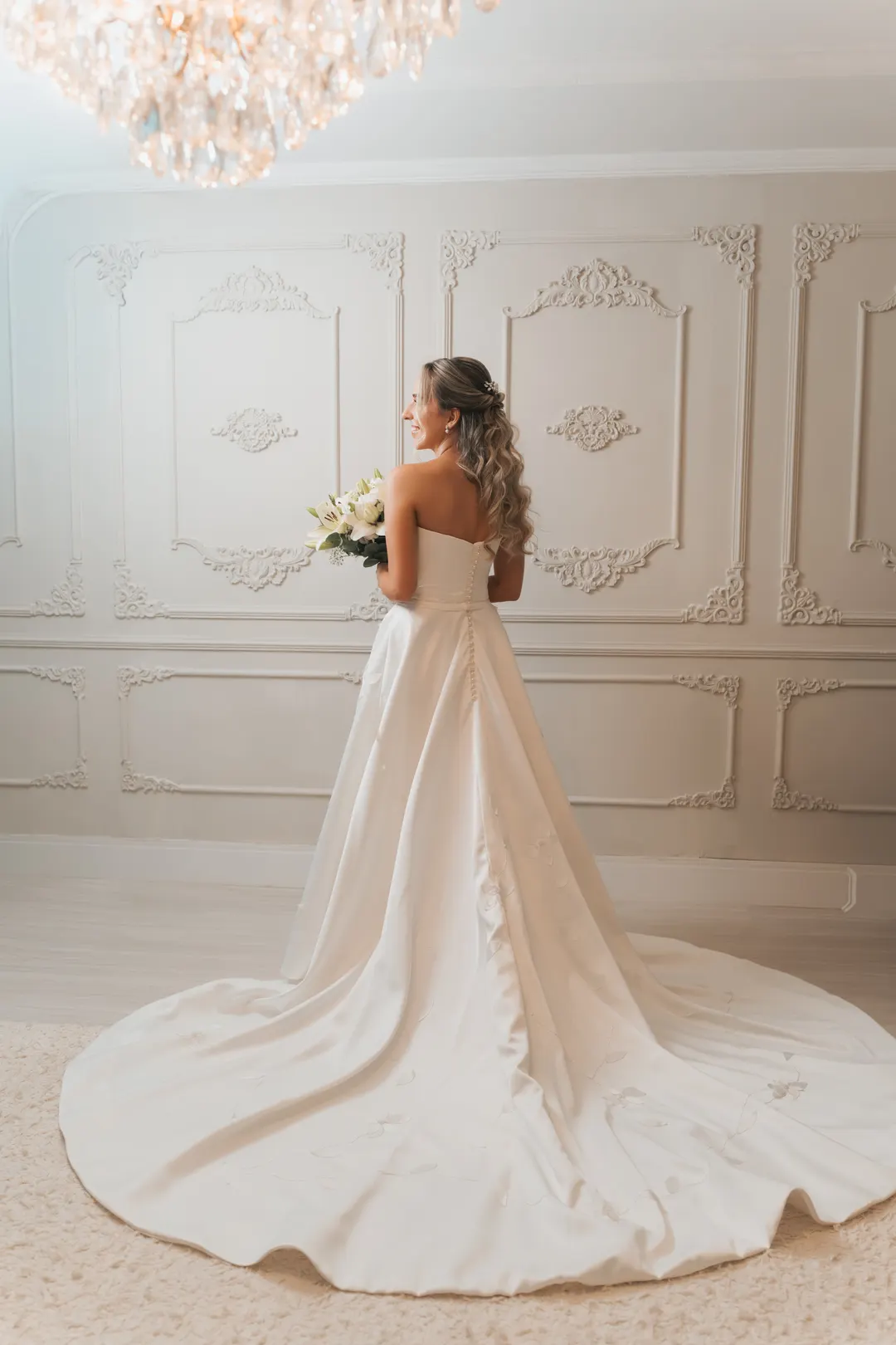 Noiva de costas em uma sala clássica branca, com a cauda do vestido aberta